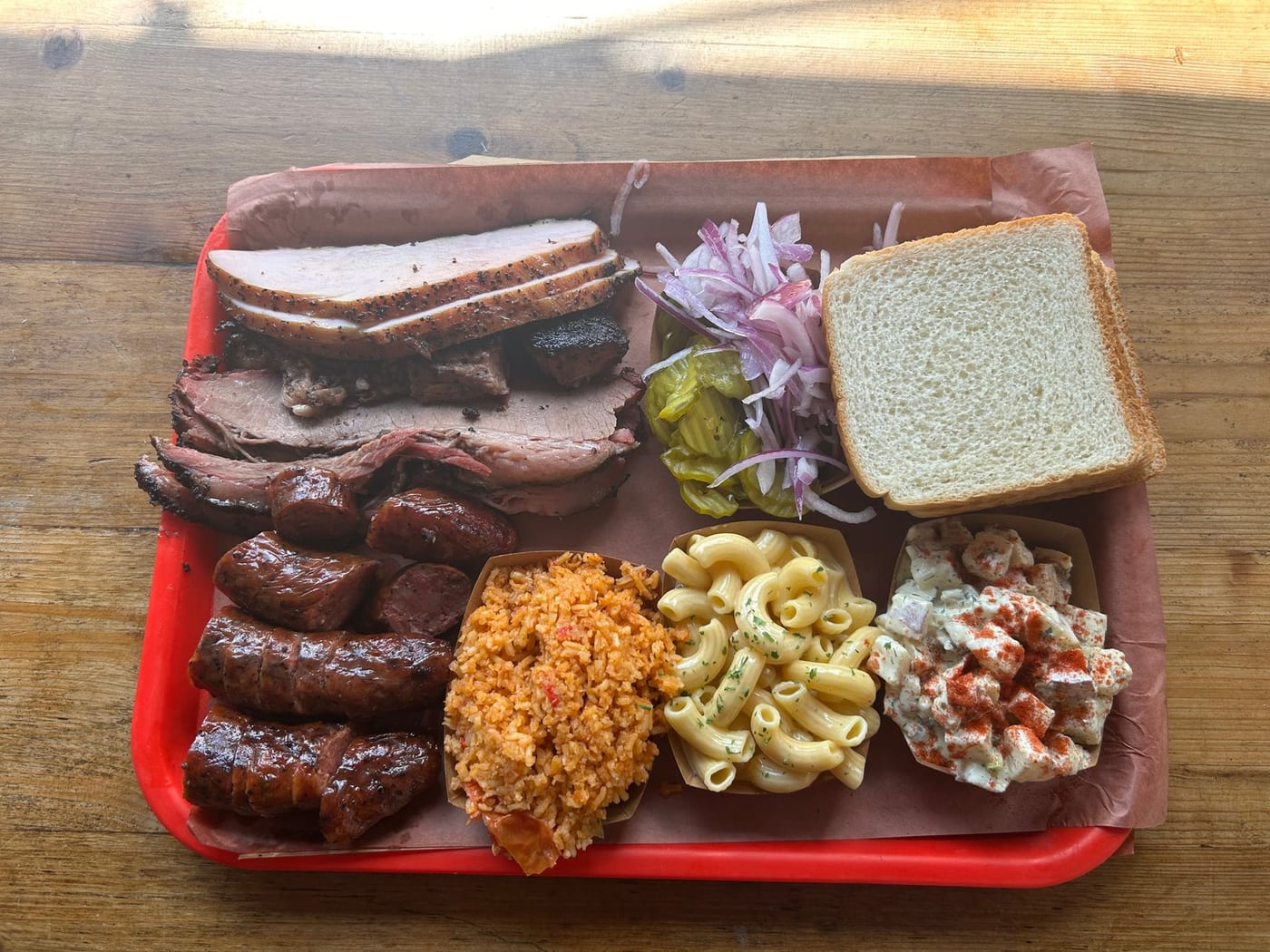 A red tray of Texas barbecue: sliced brisket, sausage links, pickles, raw onion, white bread, macaroni cheese, rice and potato salad