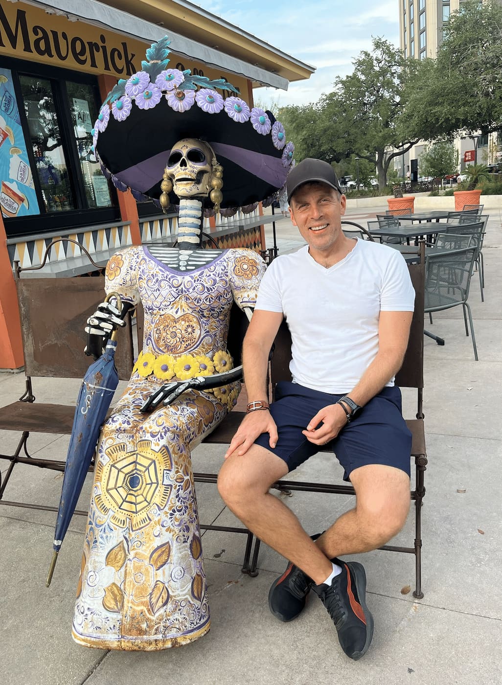 Rainer sitting on a bench beside a life-size Catrina figure in a flowered hat and painted dress, outside a cafe in San Antonio