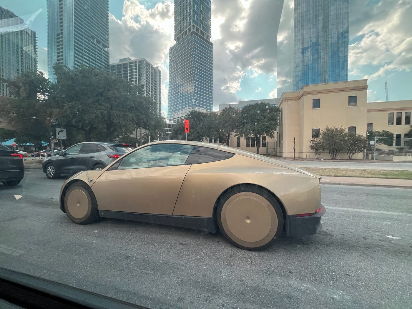 A bronze Tesla Cybercab with smooth covered wheels on a downtown Austin street, glass towers rising behind it