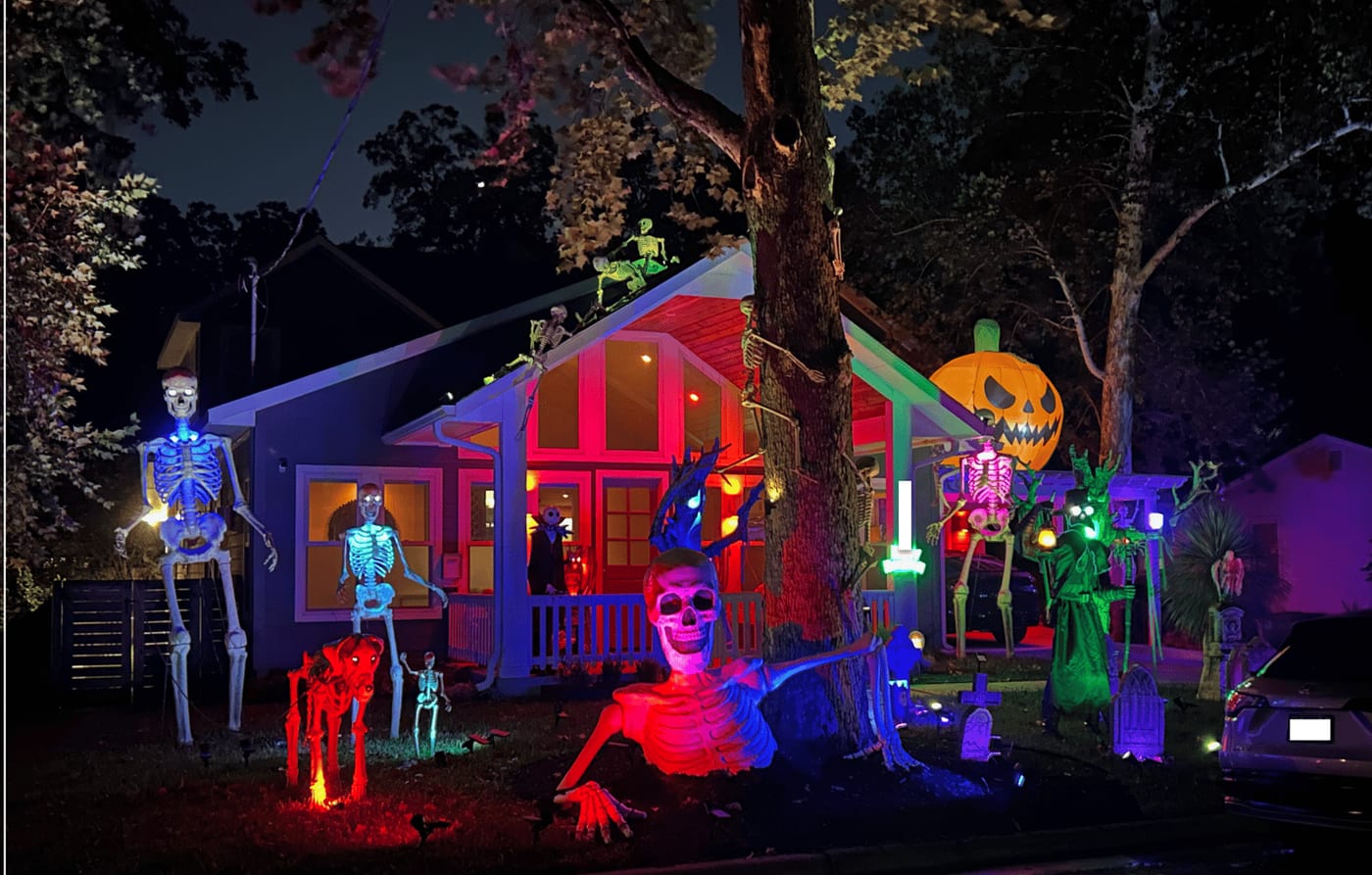 A house at night under coloured floodlights, its yard filled with giant skeletons, gravestones and a glowing pumpkin face