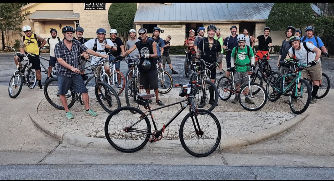 About twenty riders and their bikes gathered around a kerbed traffic island at dusk, before the Thursday ride